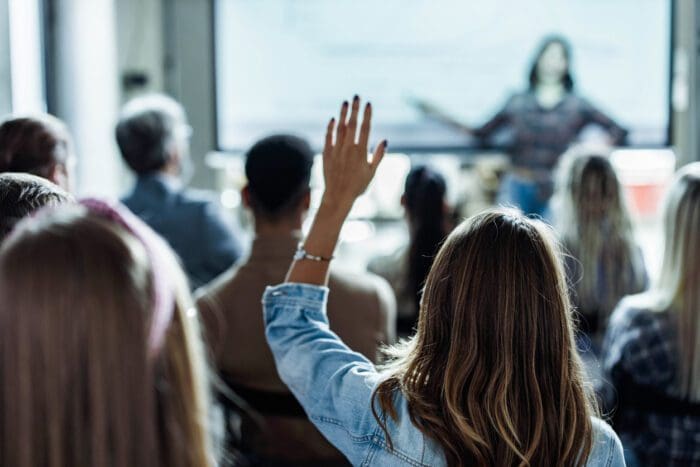 A group of people are sitting in a room facing a woman standing in front of a screen. One attendee, seen from behind in the foreground, raises their hand, presumably to ask a question or participate in the discussion. The setting appears to be a lecture or seminar, likely concluding with an FAQ session.