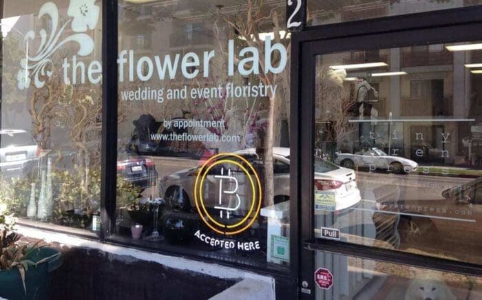 Storefront of "The Flower Lab" in Madison, WI, with a sign that reads "wedding and event floristry" and "Bitcoin accepted here." A reflection of the street and parked cars can be seen in the window. There is a potted plant at the entrance, and the door shows business hours.