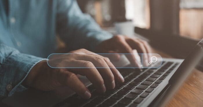 A person is typing on a laptop keyboard with a search bar superimposed in the foreground, hinting at research for a marketing framework. The scene appears to be set in a dimly lit indoor environment. The search bar includes a magnifying glass icon.