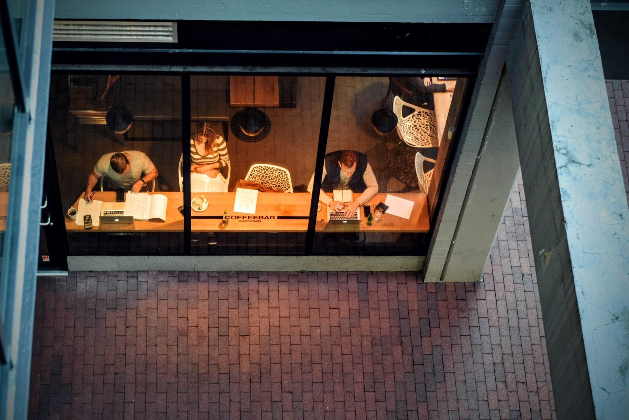 Top-down view of people sitting at a long wooden table inside a coffee shop, seen through a large window. They are reading and working on laptops with a warm light illuminating the interior, creating an ideal spot for marketing your small business. The outdoor area is paved with red bricks.