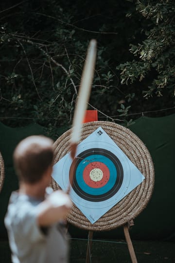 A person aiming a bow and arrow at a target placed against a green backdrop, showcasing rings with the center in red. Surrounding foliage displays asymmetric growth, adding to the scene's natural ambiance.