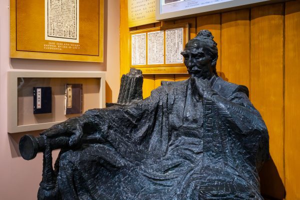 A bronze statue of a seated man deep in thought, with detailed, flowing robes and a knotted hairstyle, is displayed in a museum setting in Madison WI. The background features framed Chinese calligraphy and text, creating an academic atmosphere.