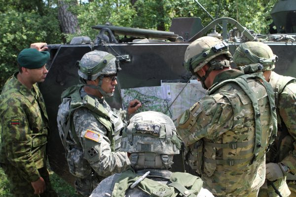 Several soldiers, dressed in camouflage uniforms and helmets, gathered around a map discussing strategy. They are outdoors near a military vehicle and surrounded by greenery. One soldier points at the map while others attentively observe, as if coordinating an asymmetric marketing campaign for a Madison WI agency.