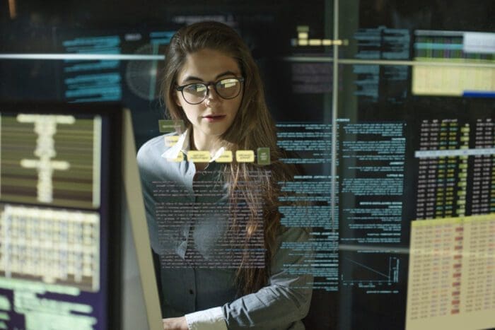 A person with long hair and glasses is sitting in front of multiple computer screens filled with code and data, likely immersed in data science tasks. The environment appears to be a tech-focused office or lab, with a futuristic, digital interface reflected over the scene.