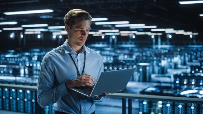 A man in a blue shirt stands in a data center, looking at a laptop. Rows of server racks with blue lights are visible in the background, with overhead lighting casting a blue hue over the large, industrial space. Focused on his work in data science, he reflects on solutions for small business challenges.