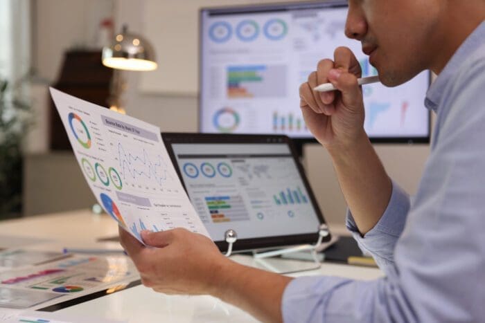 A person sitting at a desk, holding and analyzing a printed financial report with charts and graphs. In the background, a computer monitor and a laptop display additional charts and graphs, indicating data analysis and review crucial for small business decision-making through data science.