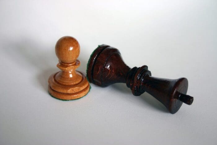 A wooden chess pawn and a fallen wooden chess king on a white surface evoke the strategy of a growth marketing agency. The light-colored, smooth-finished pawn stands tall, while the dark-colored king lies on its side, symbolizing successful checkmate tactics.