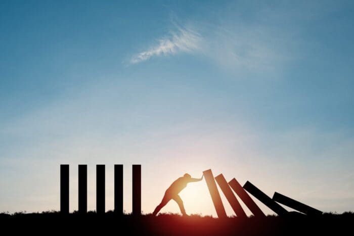 A silhouette of a person using their body to push a large rectangular block, causing a series of vertical blocks to topple like dominoes, symbolizes the cascading impact of a growth marketing agency's efforts against a bright sky and setting sun in the background.