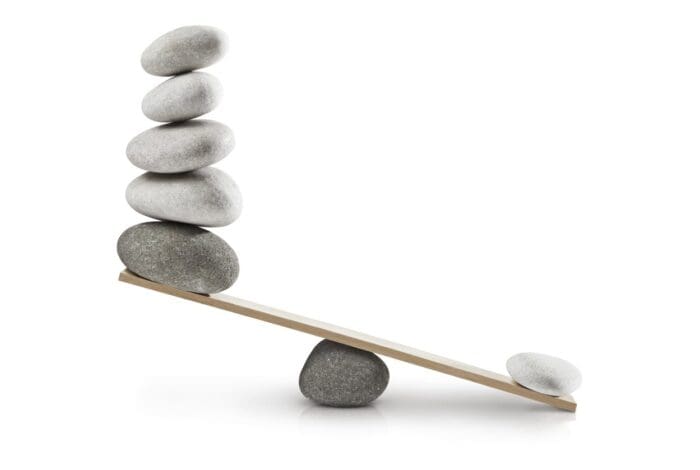 A seesaw with six smooth, grey stones stacked on the higher end, balanced by a large stone and a small stone on the lower end. The seesaw plank is supported by another stone in the center, creating an imbalance. This artistic setup speaks to guerrilla marketing tactics. White background.