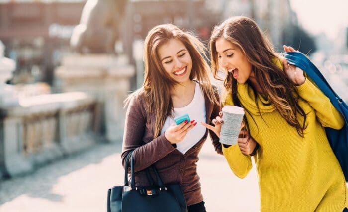 Two young women stand outdoors, smiling and looking excitedly at a smartphone one of them is holding. One woman is wearing a yellow sweater and carrying a coffee cup, while the other is dressed in a brown jacket and holding a black handbag. Their energy suggests they just stumbled upon an engaging guerrilla marketing campaign.