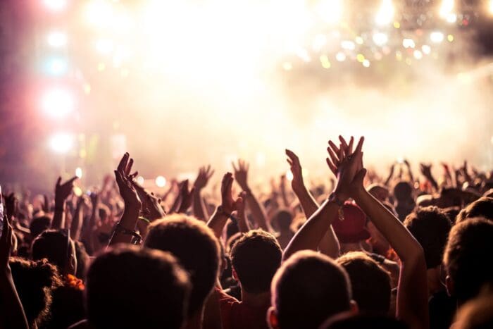 A crowd of people at an outdoor concert, raising their hands and clapping, with a brightly lit stage in the background. The atmosphere is energetic with colorful lights and some smoke, creating a vibrant and lively ambiance—perfect for guerrilla marketing strategies to engage the audience.