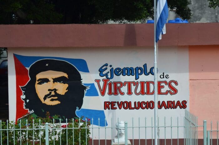 A mural on an exterior wall features a portrait of Che Guevara set against a Cuban flag. The text reads "Ejemplo de virtudes revolucionarias" (Example of revolutionary virtues). A metal fence is visible in the foreground, blending elements of guerrilla marketing with its bold political message.