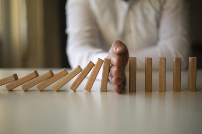 A person wearing a white long-sleeved shirt stops a sequence of falling wooden dominoes with their hand on a light-colored surface, illustrating the concept of guerrilla marketing. The dominoes on the left are mid-fall, while those on the right remain upright. The background is softly blurred.