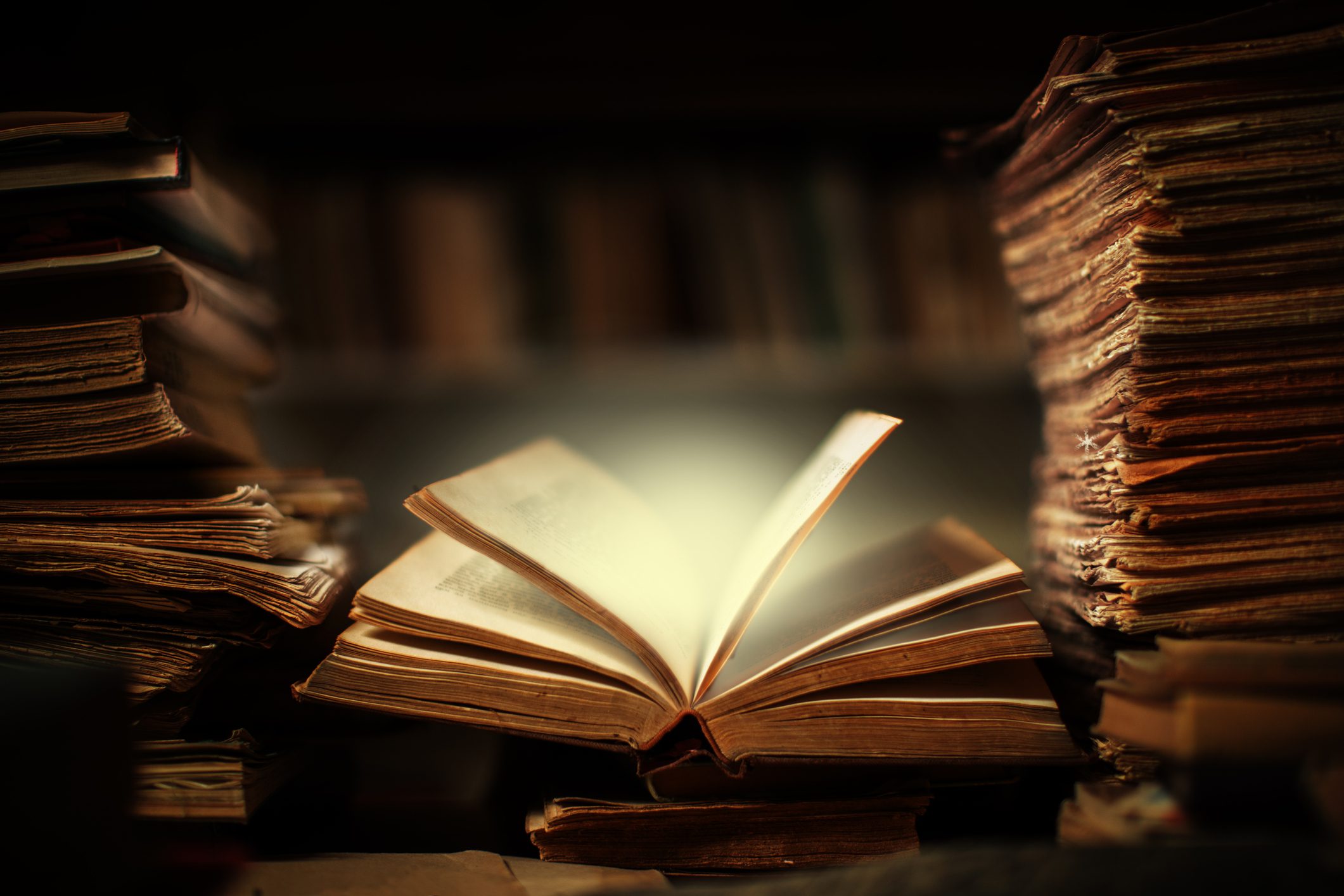 A dimly lit scene shows an open old book in the center, illuminated as if glowing. Surrounding it are tall stacks of weathered, aged books, alluding to the purpose of business and giving a sense of history and mystery. The background is blurred, making the book the focal point.