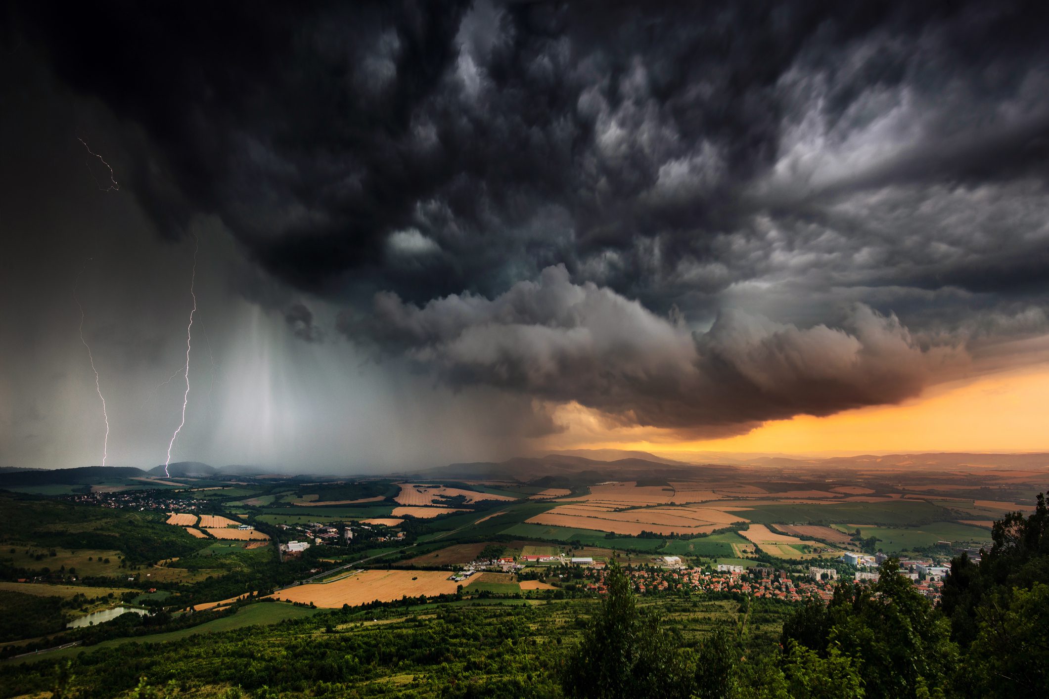 A panoramic view of a countryside under a dramatic stormy sky. Dark, thick clouds dominate the scene, with a bolt of lightning striking in the distance. A small village and fields with various shades of green and gold lie below, illuminated by a fading sunset—nature's emergency plans unfolding above.