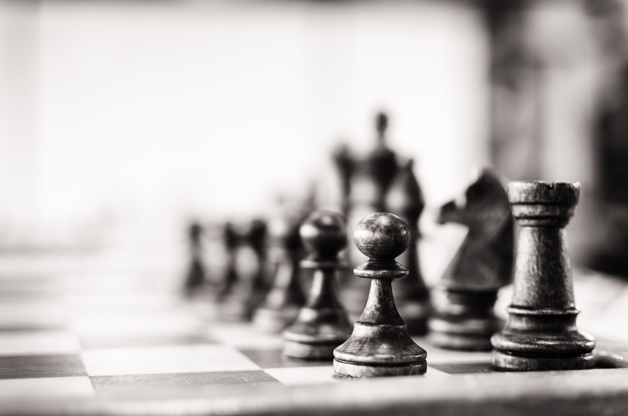 Black and white close-up image of a chessboard with focus on the black pieces, including pawns, a knight, and a rook. The background is blurred, adding depth to the portrayal of strategy development akin to intense deliberations at a conference.