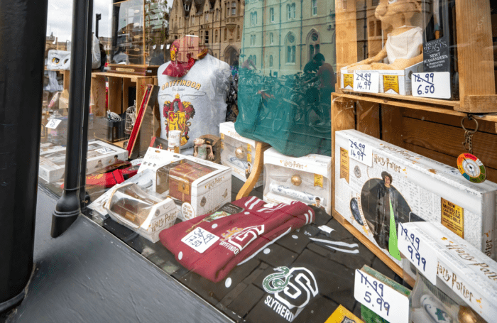 A store display features various Harry Potter merchandise, including Slytherin robes, Gryffindor sweaters, and collectible wands in boxes. A Harry Potter action figure and a selection of themed socks also adorn the shelves. Price tags and promotional signs remind shoppers that fortune favors the prepared.