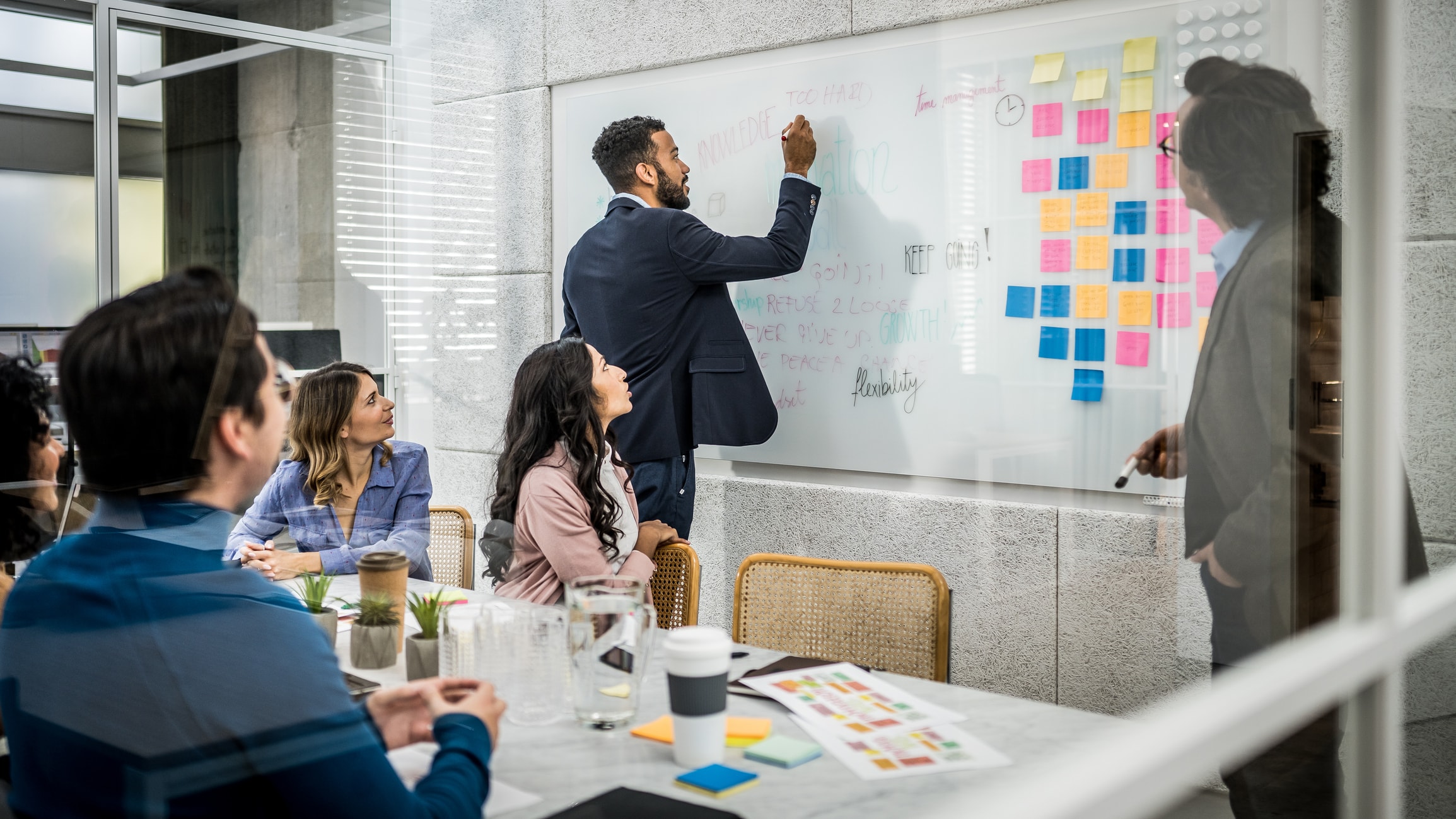 A diverse group of people in a modern office setting are collaborating on a project. One person is writing on a whiteboard filled with diagrams, guiding the team, while another points to colorful sticky notes. Others are seated around the table, engaging in discussion as if strategizing a business wargame.