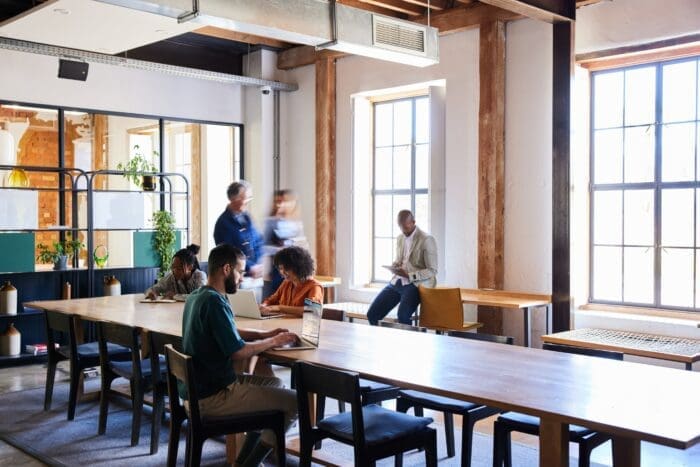 A spacious, modern office with large windows and wooden beams. Four people are working: one on a laptop at a long table, two in conversation, possibly strategizing for a business wargame, and one sitting by the window with a notebook. Indoor plants and natural light guide the creation of a bright, inviting ambiance.