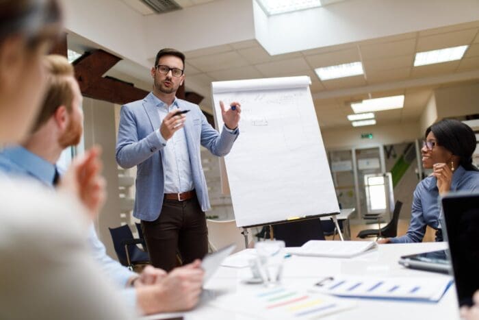 A man wearing glasses and a blue blazer is standing and speaking in front of a flip chart, guiding the team through the meeting. Three people are seated around a table with documents and a laptop, attentively listening to the presenter in what appears to be an intense business wargame session in a well-lit office space.