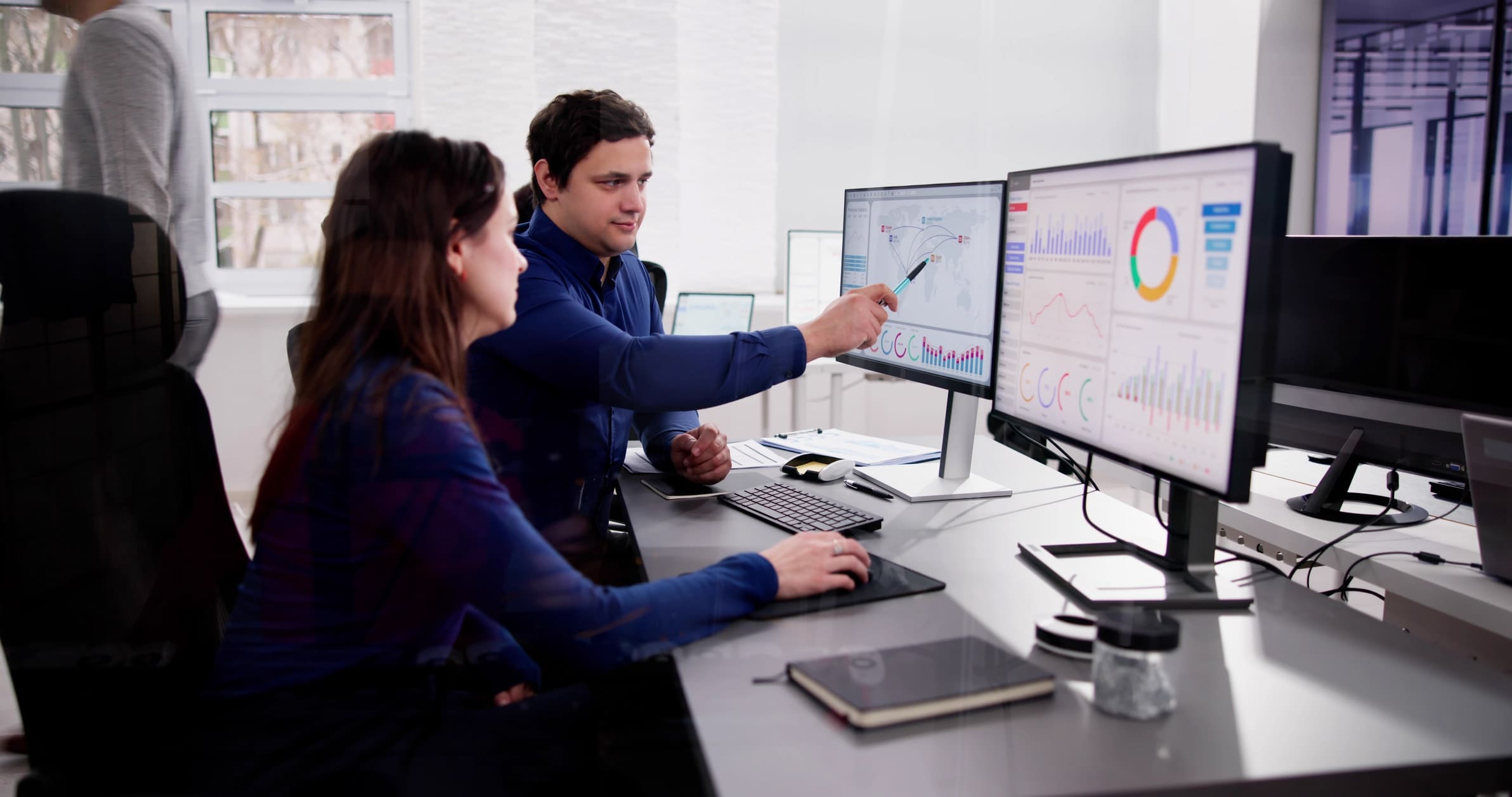 Two people are seated at a desk in an office, engaged in discussion. One person points to a computer screen displaying various charts and graphs, while the other looks on. It appears they could be immersed in a business wargame, carefully examining their strategy. The workstation includes multiple monitors, notebooks, and a coffee cup.