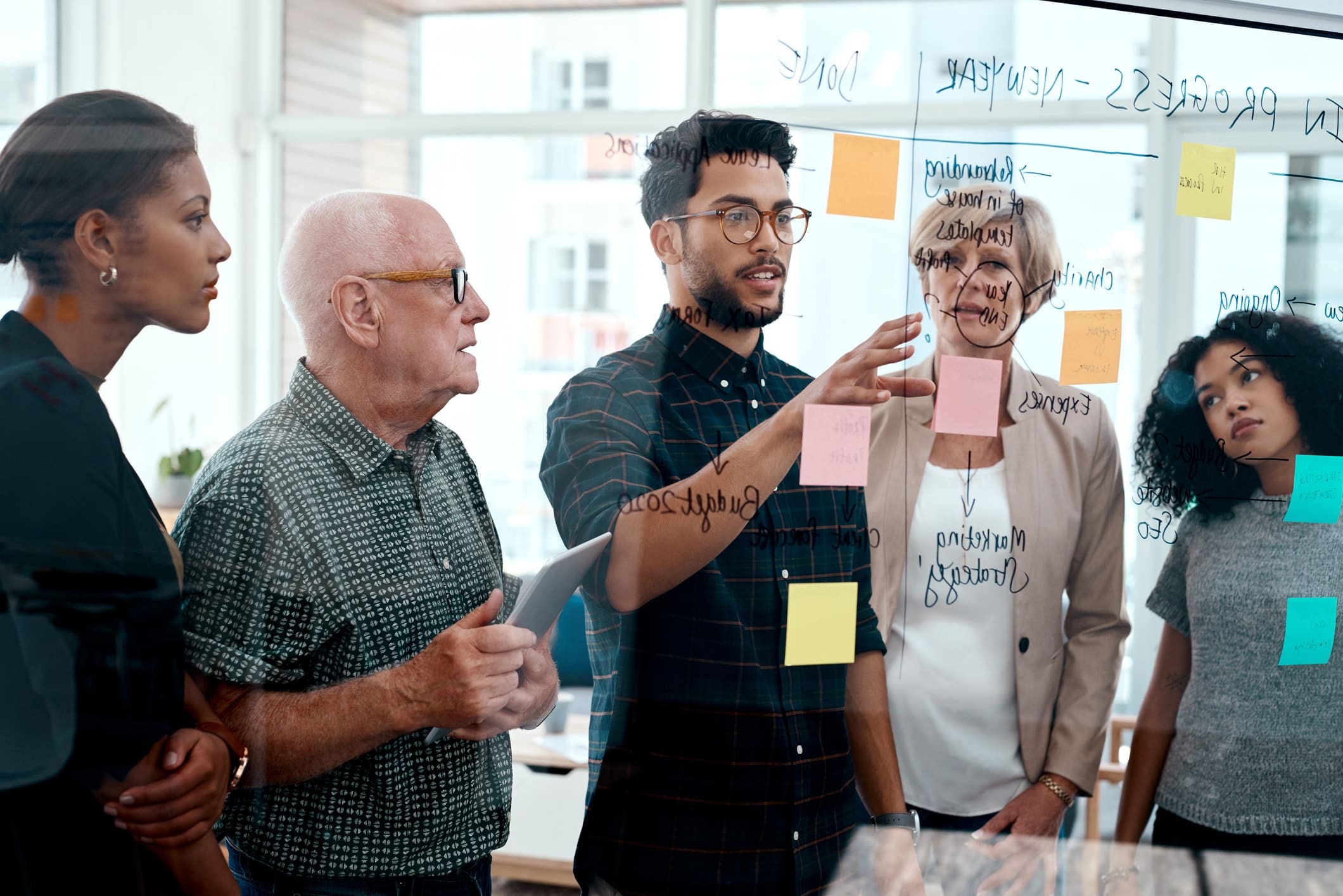 A diverse group of five people, including both men and women of different ages, are collaborating at a glass wall covered with colorful sticky notes and handwritten ideas. One person is pointing to the notes while others listen attentively, as if they were strategizing in a business wargame.