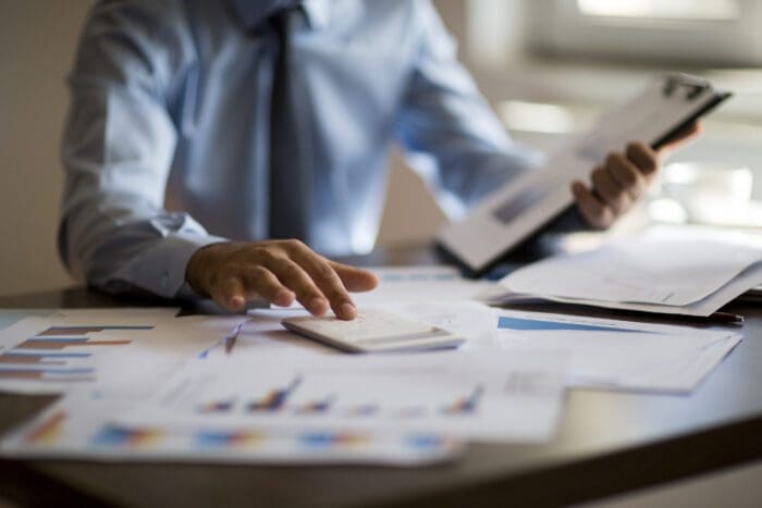 A person in a light blue shirt is seated at a desk covered with charts, graphs, and papers. They are holding a clipboard in one hand and using a calculator with the other, engaged in data analysis or competitive intelligence work.