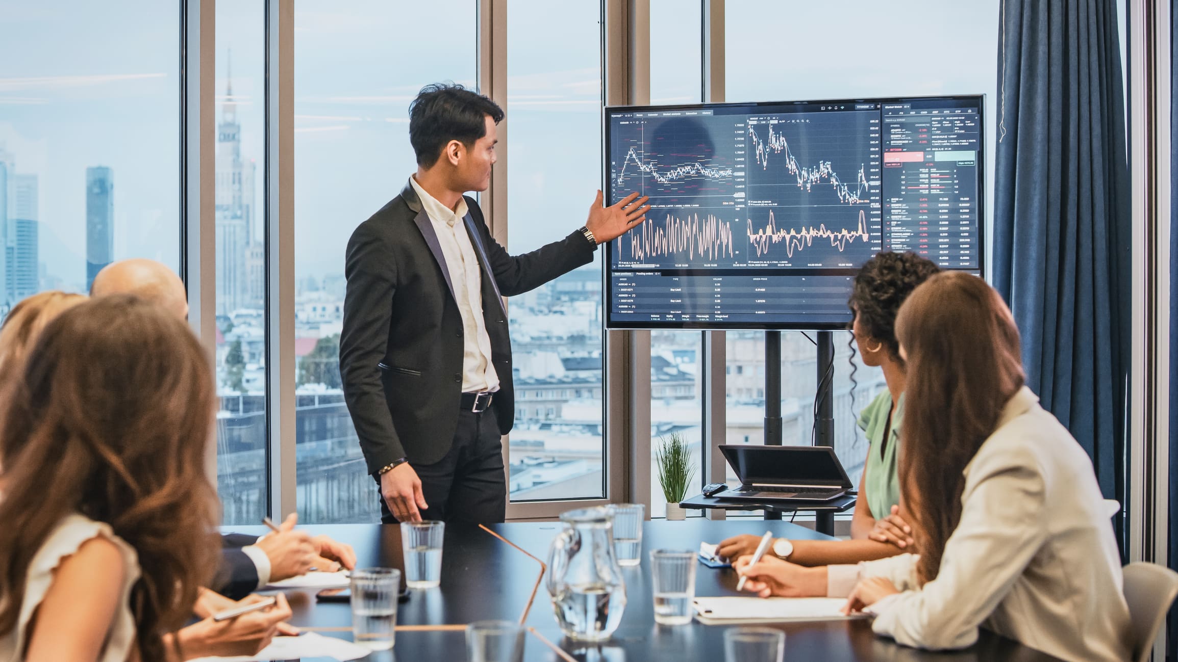 A man in a suit stands in front of a large screen displaying financial data and charts, addressing a group of people seated around a conference table. He shares insights on competitive intelligence. The room has large windows with a cityscape view outside.