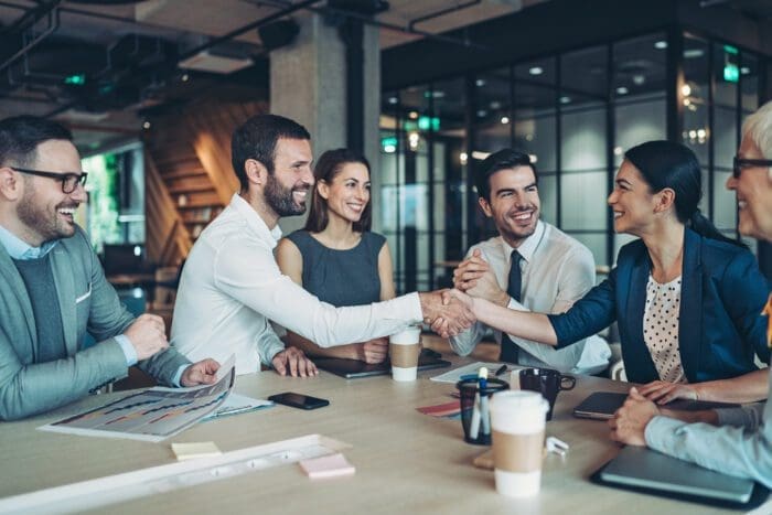 A diverse group of professionals sits around a conference table in a modern office. Two people in the middle shake hands while others smile and chat. Laptops, papers, and coffee cups are on the table, hinting at discussions centered on competitive intelligence. The atmosphere appears collaborative and positive.