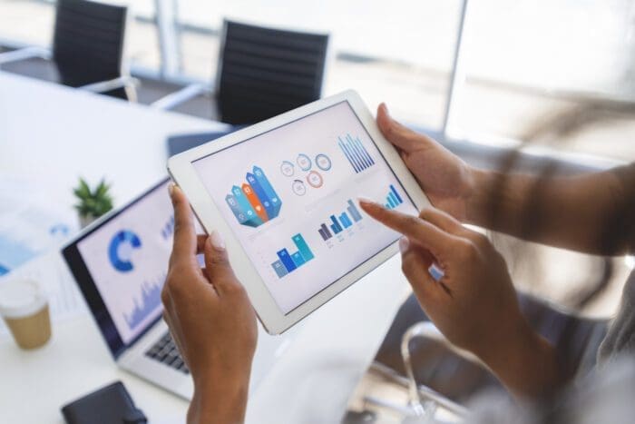 Hands holding a tablet displaying colorful business analytics graphs and charts. A blurred background shows a laptop with a similar chart, documents, and a coffee cup on a conference table, suggesting competitive intelligence gathering during a business meeting or analysis session.