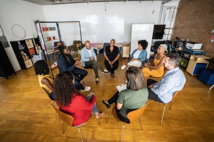 A group of nine people is seated in a circle in a brightly lit room with wooden floors. They are engaged in a discussion on competitive intelligence, with some holding papers or notebooks. Shelving, a whiteboard, and various office supplies are visible in the background.