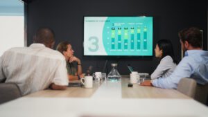 Four people sitting around a conference table in an office, attentively looking at a screen displaying a bar chart titled "Numbers of Customer". Two people have laptops open, suggesting they're gathering competitive intelligence. Coffee mugs and a water carafe are also on the table.