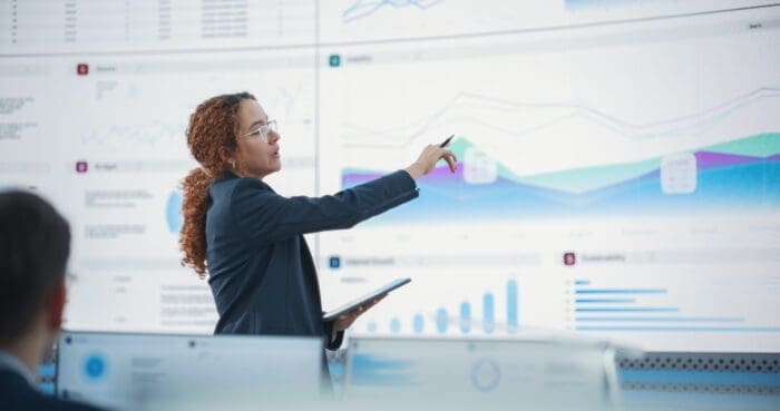 A person with curly hair, wearing glasses and business attire, points to a large digital screen displaying charts and graphs. They hold a tablet in one hand. The background shows multiple digital screens with data and visualizations, emphasizing competitive intelligence.
