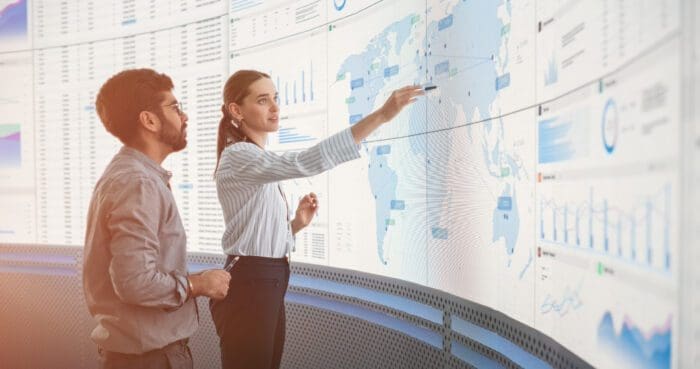 Two professionals stand in front of a large digital screen displaying various charts, graphs, and a world map. One person points at the screen while discussing the data, highlighting their collaborative analysis in uncovering competitive intelligence.