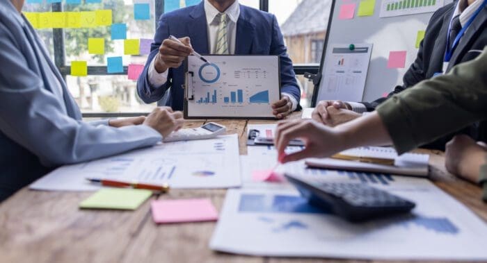 A group of business professionals is gathered around a table during a meeting. One person is holding a financial chart, pointing at a graph, while others have documents, calculators, and sticky notes essential for competitive intelligence. The background features sticky notes on a window and a whiteboard with charts.