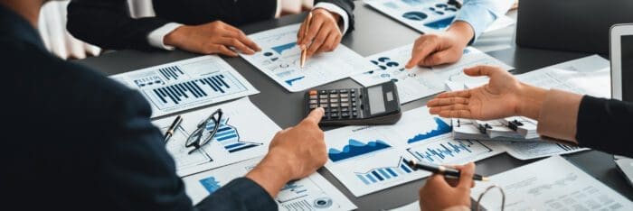 A group of people sit around a table covered with various financial documents, charts, and graphs. One person points to a calculator, while others discuss competitive intelligence strategies and gesture towards the papers. Eyeglasses, pens, and a laptop are also visible on the table.