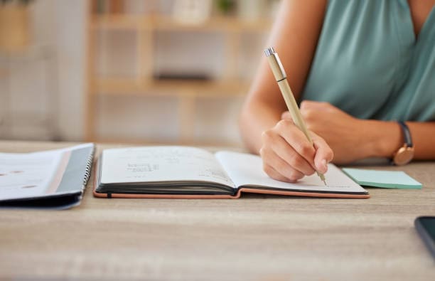 Close-up of a person's hand writing in a notebook with a pen. The individual is wearing a sleeveless top and a watch, seated at a wooden table with other documents and books. The softly blurred background highlights the peaceful workspace setting where it's clear that fortune favors the prepared.