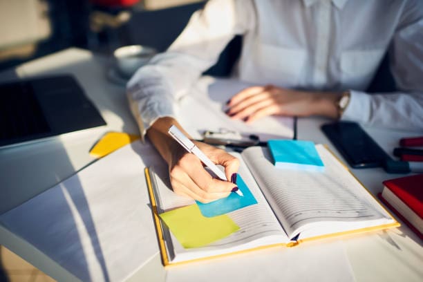 A person sits at a desk, writing on a blue sticky note with a pen. The desk is cluttered with an open notebook, a laptop, a smartphone, and scattered papers. Wearing a white shirt, they seem immersed in their tasks—after all, fortune favors the prepared. Their face is not visible in the image.