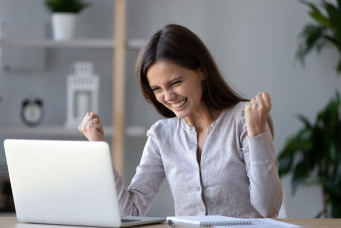 A woman wearing a light gray sweater is seated at a desk, looking at a laptop. She is smiling and clenching her fists in excitement, clearly embodying the motto "Diligence is the Mother of Good Luck." A notebook and pen are on the desk, with a potted plant and shelves in the background.