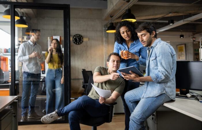 A group of colleagues collaborates in a modern office. Three people focus on a tablet and documents at a desk, immersed in strategic planning, while two others chat in the background by a glass door. The setting includes industrial decor with exposed concrete and ceiling ducts.