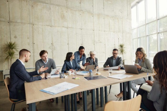 A diverse group of eight people, embodying Sun Tzu's strategic principles, are seated and standing around a long wooden table in a modern office. Engrossed in discussion, some use laptops while others consult documents or tablets. Large windows and concrete walls frame the scene in the background.