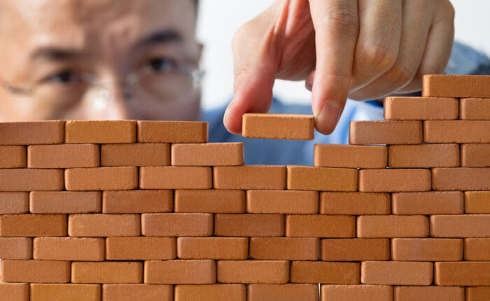 A person wearing glasses places a small brick into a gap on top of a wall made of miniature bricks, focusing intently on the task. The image symbolizes construction, building, or problem-solving, reflecting the idea that diligence is the mother of good luck.