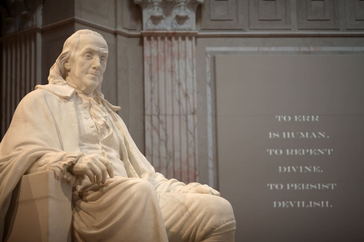 A marble statue of Benjamin Franklin seated and wearing historical attire is prominently displayed indoors. To the right, a wall features the inscription: "To err is human, to repent divine, to persist devilish." The setting is grand with ornate columns in the background, a testament to "Diligence is the Mother of Good Luck.