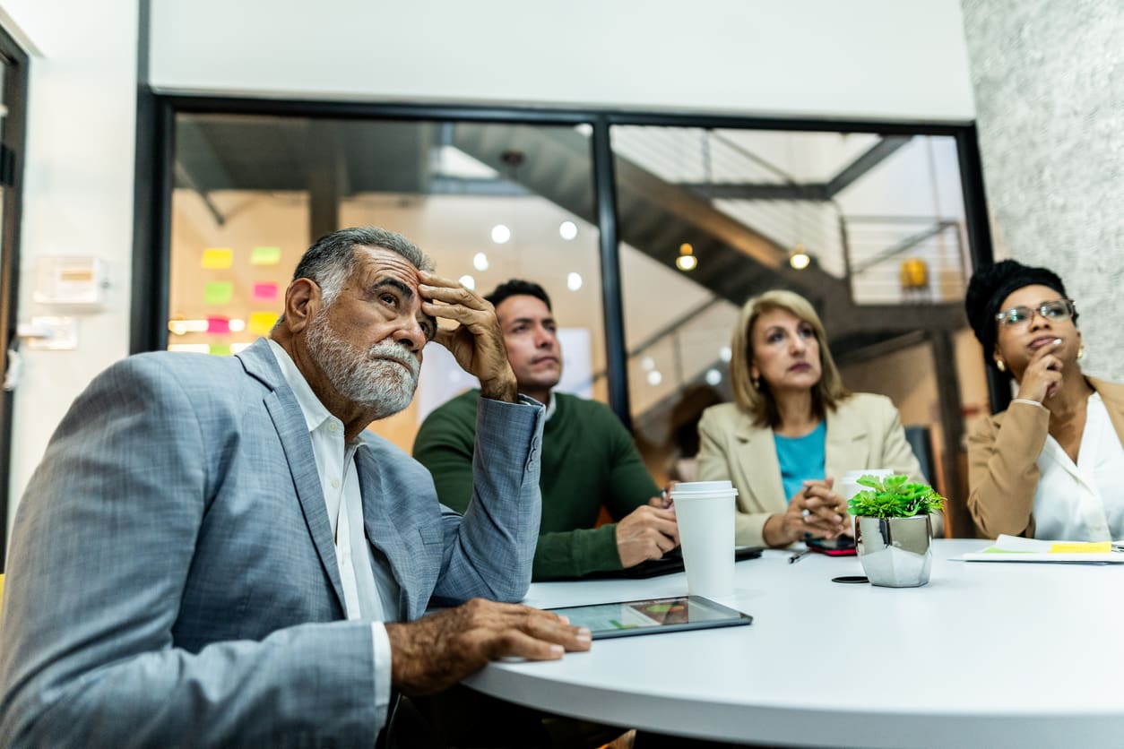 A group of four people, three men and one woman, are gathered around a table in a modern office. They appear to be attentively listening to a presentation on crisis management. One person is holding a tablet, while another has a cup of coffee in front of them. A potted plant sits on the table.