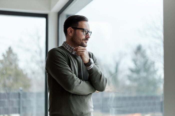 A man with glasses and a beard stands near a large window, looking thoughtfully into the distance with his hand resting on his chin, reminiscent of a modern-day Sun Tzu. He is wearing a green sweater over a checked shirt. Trees and an overcast sky are visible outside the window.