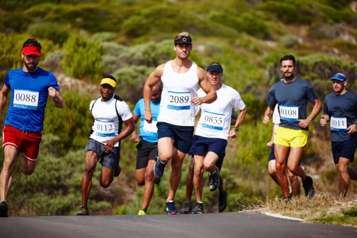 A group of runners wearing numbered bibs participates in an outdoor race on a road surrounded by greenery. The runners, dressed in athletic attire, are captured mid-stride with determined expressions, embodying the saying "Diligence is the Mother of Good Luck." The sky is clear, and the overall scene is vibrant and dynamic.