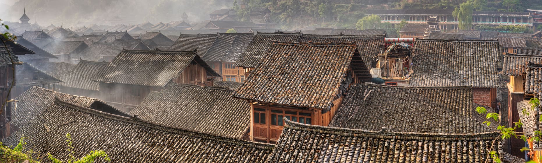 Panoramic view of a traditional village with numerous closely packed wooden houses featuring distinctive tiled roofs. The scene, reminiscent of Sun Tzu’s era, is shrouded with a light mist, adding an atmospheric touch to the historic architecture. Greenery can be seen in the background.