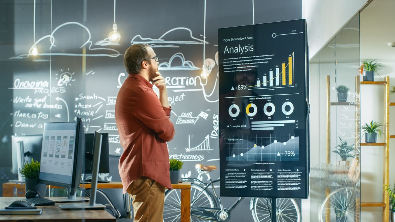 A man stands in front of a large data screen displaying various graphs and charts. He appears deep in thought, holding his chin as he considers the results of content analysis. Behind him, a glass wall is covered with handwritten notes and diagrams, and there is a desk with computers and office equipment.