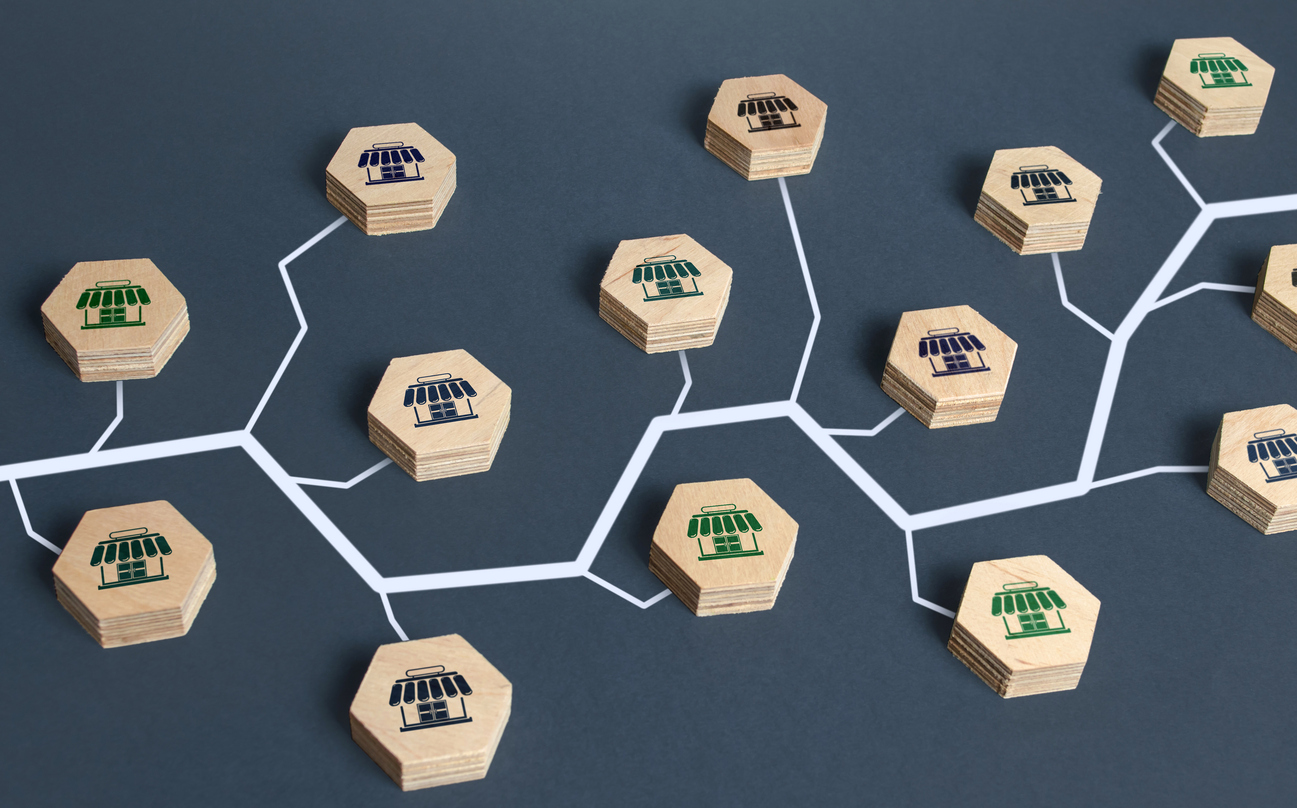 An abstract representation of a franchise network or organizational structure using wooden hexagons with store icons interconnected by white lines on a dark background, illustrating the importance of franchise SEO. The hexagons feature blue, green, and black storefront symbols.