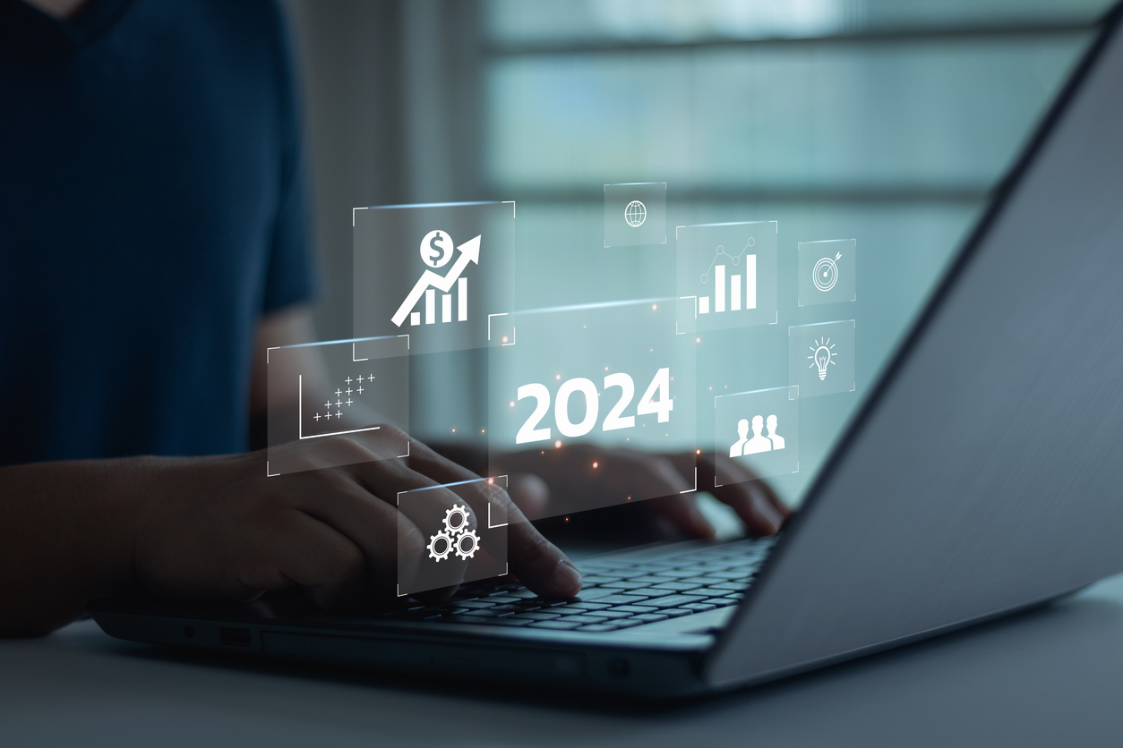 A person typing on a laptop with glowing digital icons hovering around, indicating financial growth, analytics, graphs, and team collaboration. The central glowing text reads "2024," symbolizing future progress and projections in the marketing budget.